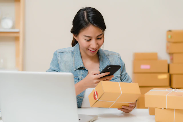 Une commerçante vérifie une commande sur son téléphone, entourée de colis